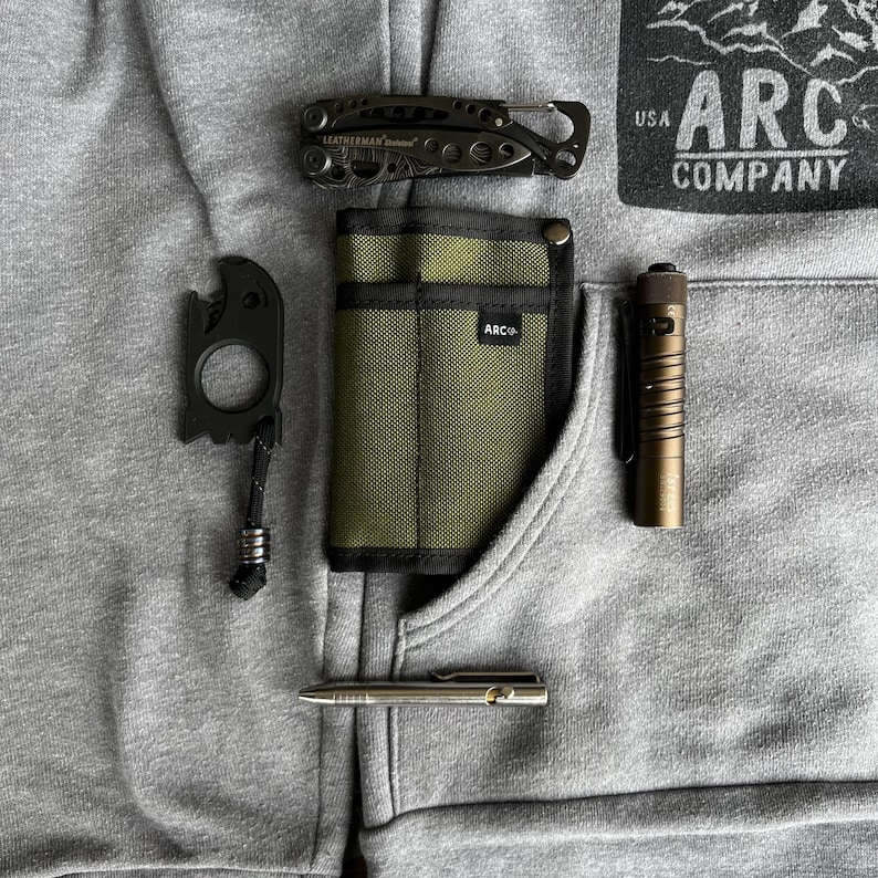 May include: A gray hoodie with a black and green pocket organizer, a black multi-tool, a black bottle opener, a silver pen, and a brown flashlight. The hoodie has a patch on the front that says "USA ARC COMPANY".