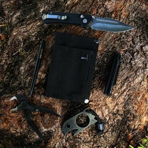 May include: A black fabric pouch with a pocket and the word "ARCO" printed on it.  A black folding knife, a black pen, a black flashlight, a black multi-tool with a ring, and a black multi-tool with a hook are also pictured.