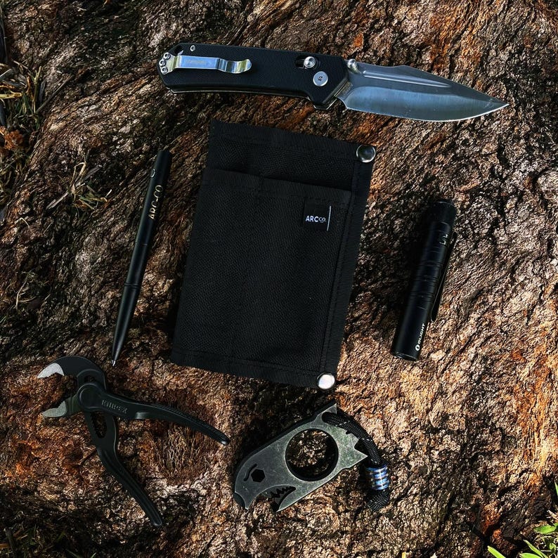 May include: A black fabric pouch with a pocket and the word "ARCO" printed on it.  A black folding knife, a black pen, a black flashlight, a black multi-tool with a ring, and a black multi-tool with a hook are also pictured.