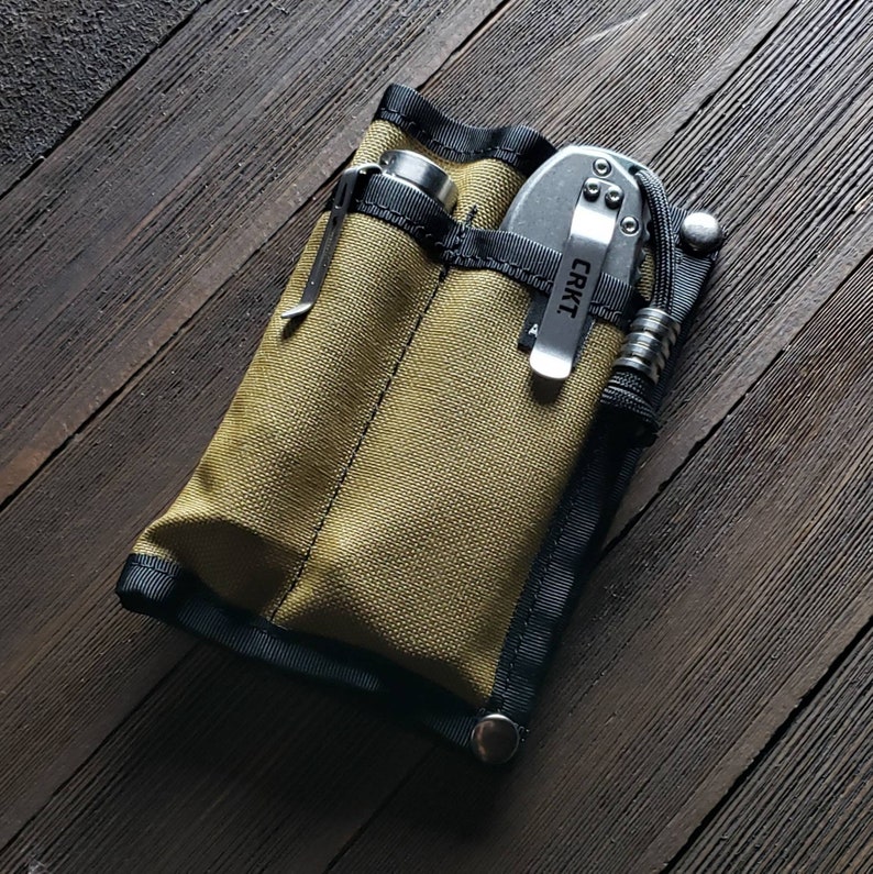 May include: A khaki-colored utility pouch with black trim and a zipper closure. The pouch has two pockets, one for a flashlight and one for a folding knife. The knife is a silver CRKT brand knife with a black handle.
