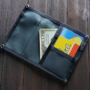 May include: A black fabric wallet with a zipper closure and a flap. The wallet has a billfold, a card slot, and a small pocket. The wallet is open and contains a $20 bill and a yellow card with the word "Card" printed on it.