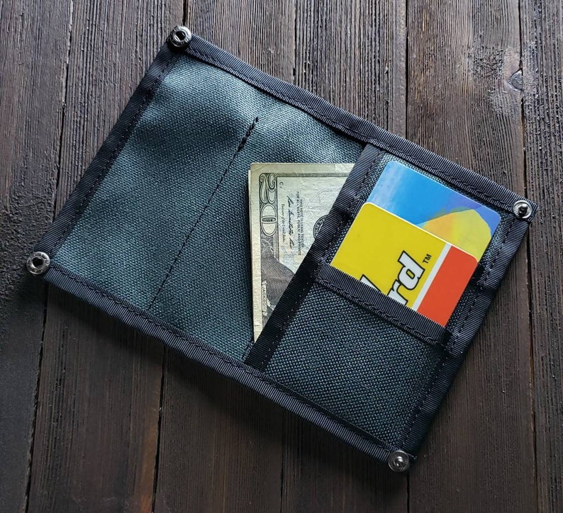 May include: A black fabric wallet with a zipper closure and a flap. The wallet has a billfold, a card slot, and a small pocket. The wallet is open and contains a $20 bill and a yellow card with the word "Card" printed on it.