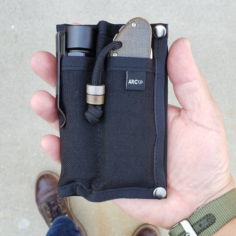 May include: A black nylon pouch with a zipper closure and a snap button. The pouch has two pockets, one for a flashlight and one for a knife. The pouch has the text "ARC CO." printed on it.