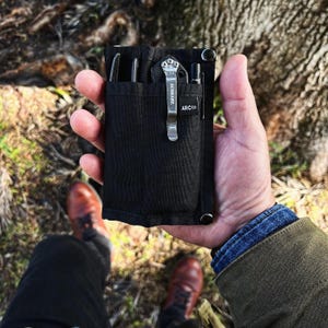 May include: A black nylon pouch with a zipper closure and multiple pockets. The pouch is holding a small pocket knife, a pen, and a multi-tool. The pouch has the text "ARC'CO" and "SCHRADE" printed on it.