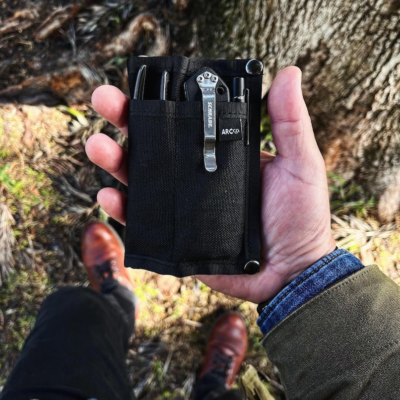 May include: A black nylon pouch with a zipper closure and multiple pockets. The pouch is holding a small pocket knife, a pen, and a multi-tool. The pouch has the text "ARC'CO" and "SCHRADE" printed on it.