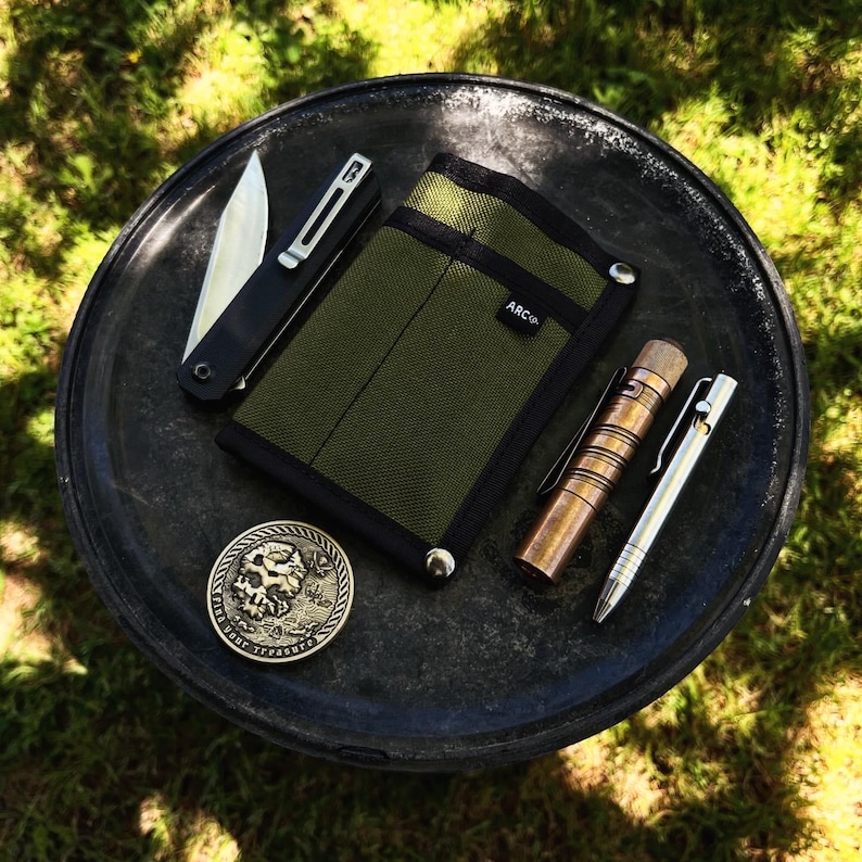 May include: A green canvas pocket organizer with a black leather trim and the word "ARC'" printed on it. The organizer is on a black metal tray with a silver pocket knife, a copper flashlight, and a silver pen.