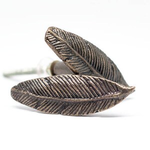 May include: Two bronze-coloured feather-shaped cabinet knobs. The knobs have detailed veining and are set against a white background. The hardware is designed for furniture and adds a decorative touch.