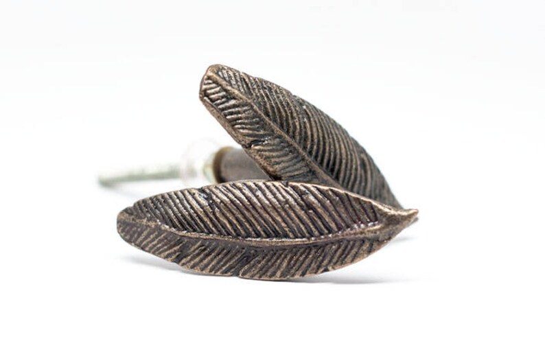 May include: Two bronze-coloured feather-shaped cabinet knobs. The knobs have detailed veining and are set against a white background. The hardware is designed for furniture and adds a decorative touch.