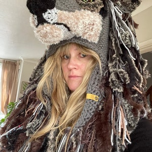 May include: A grey crocheted wolf hat with a long, shaggy mane of brown, grey, and white yarn. The hat has a realistic wolf face with black eyes and a black nose.