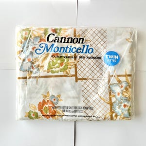 May include: A package of a twin-sized fitted sheet in a floral pattern with a white background. The sheet is made by Cannon Monticello and is a no-iron percale bed fashion. The package says "Twin Fitted" and "50% Polyester + 50% Combed Cotton (Coton) Made in U.S.A."