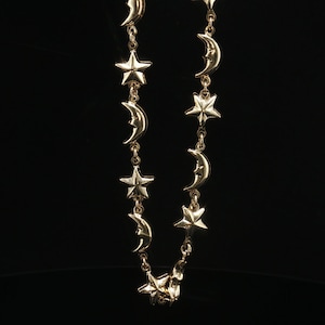May include: A gold-toned chain necklace featuring alternating polished metal charms shaped like five-pointed stars and crescent moons. The individual charms are linked together in a continuous vertical arrangement with small circular jump rings.