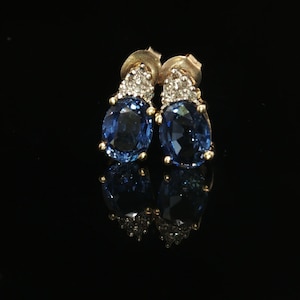 May include: A pair of stud earrings featuring oval-cut deep blue gemstones set in gold. Each earring is accented with a small cluster of clear, sparkling stones at the top, secured with gold earring backs against a dark background.