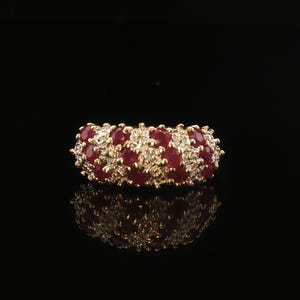 Op de afbeelding: Een gouden ring met een cluster van ronde, dieprode robijnen afgewisseld met kleine, heldere, fonkelende diamanten. De stenen zijn in een dichte, meerrijige opstelling over de bovenkant van de ring gezet, wat zorgt voor een textuurrijk en schitterend oppervlak.
