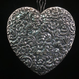 May include: A heart-shaped silver pendant featuring an intricate, raised relief pattern of various flowers and swirling scrollwork. The metallic surface has a polished finish with dark recessed areas that highlight the detailed floral design.