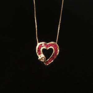 May include: A gold necklace featuring a heart-shaped pendant. The pendant is outlined with a row of round, deep red rubies and contains a smaller, solid gold heart accent at the bottom left corner, suspended from a delicate gold box chain.