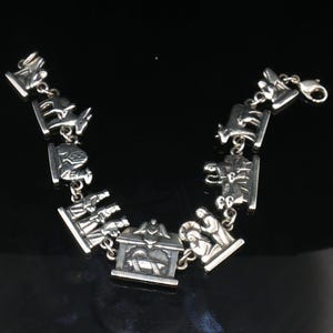 Puede incluir: Una pulsera de dijes en tono plateado con una serie de eslabones metálicos que representan una escena de natividad. Los dijes incluyen figuras de animales, tres reyes magos, un pesebre central con un ángel encima y dos figuras arrodilladas.