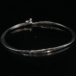 Peut inclure: Un collier torque en argent sterling poli avec une bande effilée et lisse formant un cercle. La pièce présente un petit mécanisme de fermeture de type crochet intégré sur le dessus, avec une finition métallique très réfléchissante.