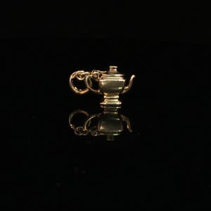 May include: A small gold-toned metal charm shaped like a traditional teapot with a curved spout and a lid. The charm features a circular jump ring attached to the handle side, allowing it to be suspended from a chain or bracelet for jewelry use.