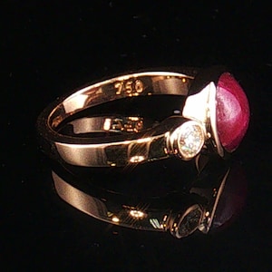 May include: A gold ring featuring a smooth, cabochon-cut deep red gemstone and a small, round, clear accent stone. The inner band is clearly stamped with the number 750, indicating the gold purity, and the metal has a polished, reflective finish.