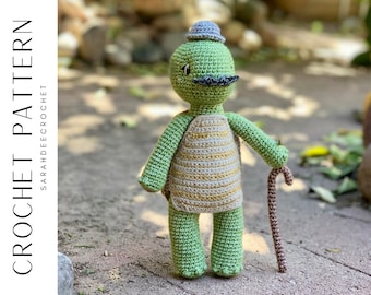 May include: A crocheted green turtle wearing a striped vest and a purple hat. The turtle has a brown mustache and is holding a brown walking stick.