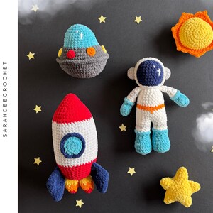 Pode incluir: Conjunto de padrões de crochê amigurumi com um foguete vermelho e branco, uma nave espacial azul e branca, um astronauta branco com cinto laranja e um sol amarelo. O padrão está disponível para download.