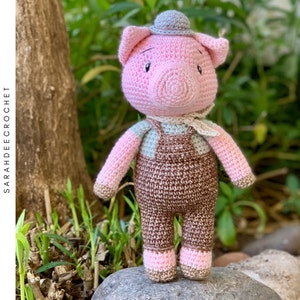 May include: A crocheted pink pig wearing a brown overall and a gray hat. The pig is standing on a rock.