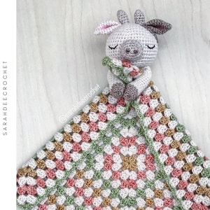 May include: A crocheted baby blanket with a crocheted animal toy attached. The blanket is made with a multi-colored granny square pattern in shades of pink, green, white, and brown. The animal toy is a gray and white cow with closed eyes and a brown nose.