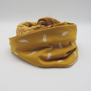 May include: A mustard yellow infinity scarf featuring a white feather print on the exterior and a white and mustard yellow striped pattern on the interior. The fabric is folded to show both the feather-patterned side and the striped lining.
