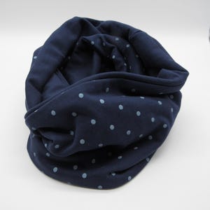 May include: A dark navy blue infinity scarf featuring a repeating pattern of small, light blue polka dots. The fabric has a soft, textured appearance and is arranged in a loose, circular loop, showcasing the interior and exterior folds.