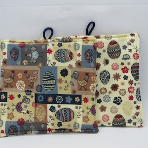 Könnte beinhalten: Zwei gelbe und braune Topflappen mit Patchwork-Muster mit Hühnern und Eiern. Die Topflappen haben einen blauen Rand und eine Schlaufe zum Aufhängen.