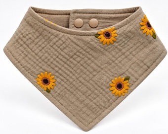 May include: A triangular bandana bib in a muted taupe color with a textured, multi-layered fabric finish. The bib features embroidered sunflower motifs with yellow petals and dark centers, and it secures with two matching taupe plastic snap buttons.