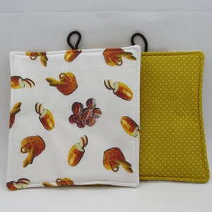 Könnte beinhalten: Zwei Topflappen in Weiß und Gelb. Ein Topflappen hat einen weißen Hintergrund mit einem Muster aus braunem Brot und einem braunen Krug. Der andere Topflappen hat einen gelben Hintergrund mit weißen Punkten.