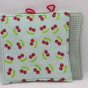 Puede incluir: Dos agarraderas para ollas, una con fondo azul claro y un patrón de cerezas rojas, y la otra de tejido de gofre verde claro liso.