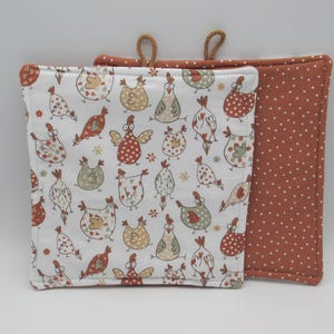 May include: Two square pot holders. One is white with a repeating pattern of cartoon chickens in various colours. The other is a solid rust colour with white polka dots. Both have a loop for hanging.