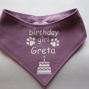 May include: Purple bandana with white text that says "birthday girl Greta" and has two paw prints and a birthday cake with a candle on top.