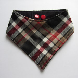 May include: A triangular dog bandana with a plaid design. The pattern includes black, red, white, and beige checks. Two red buttons secure the top. The item is likely made of fabric.