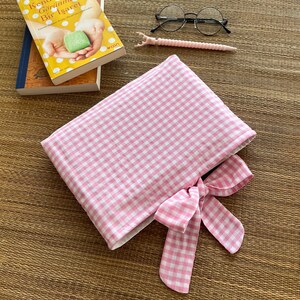 May include: A pink and white gingham book sleeve with a matching bow, next to a pair of round glasses, a pink pen, and two books. The book on top has a yellow cover with Turkish text.