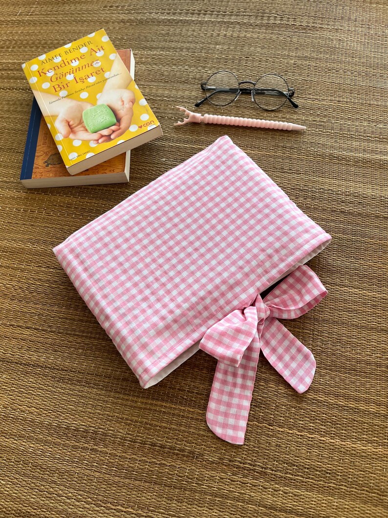 May include: A pink and white gingham book sleeve with a matching bow, next to a pair of round glasses, a pink pen, and two books. The book on top has a yellow cover with Turkish text.