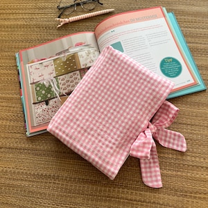 May include: A pink and white gingham fabric pouch with a bow, lying on an open book with colorful illustrations. A pair of glasses and a pink pen are also on the surface. The book's title is partially visible.