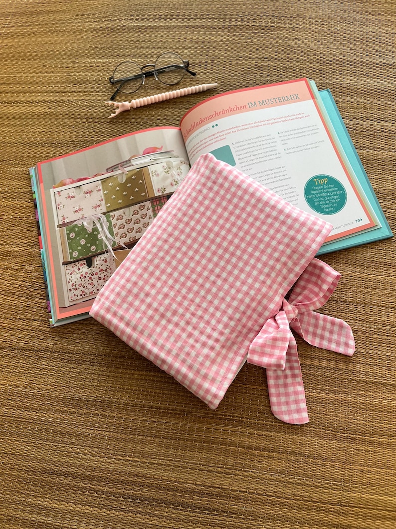 May include: A pink and white gingham fabric pouch with a bow, lying on an open book with colorful illustrations. A pair of glasses and a pink pen are also on the surface. The book's title is partially visible.
