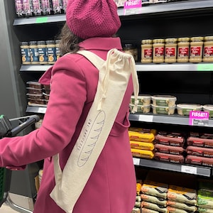May include: A person wearing a magenta coat and hat, carrying a beige baguette bag with the text "UNE BAGUETTE" and a baguette illustration. The person is in a grocery store with shelves of food items.