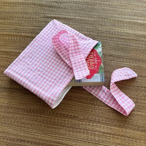 May include: A pink and white gingham book sleeve with a matching ribbon closure. The book sleeve is open, revealing a book with a red and white cover. The book and sleeve are on a woven mat.