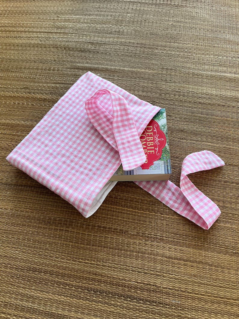 May include: A pink and white gingham book sleeve with a matching ribbon closure. The book sleeve is open, revealing a book with a red and white cover. The book and sleeve are on a woven mat.