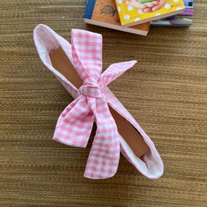 May include: A pink and white gingham fabric book sleeve with a bow closure. The interior is white, and the book sleeve is resting on a woven brown surface. Several books are in the background.