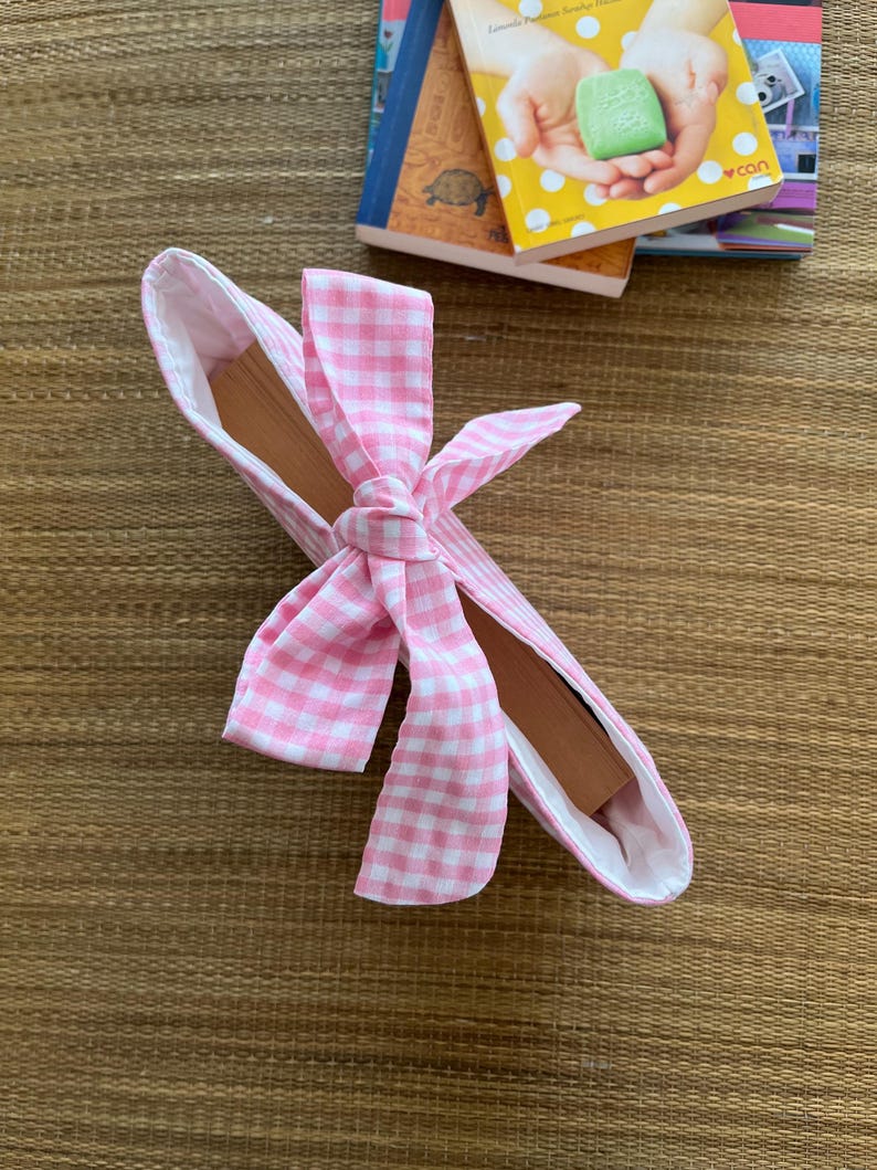 May include: A pink and white gingham fabric book sleeve with a bow closure. The interior is white, and the book sleeve is resting on a woven brown surface. Several books are in the background.