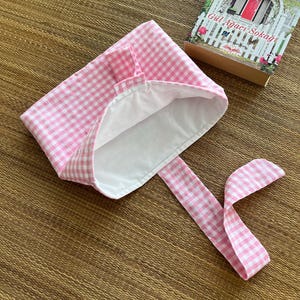 May include: A pink and white gingham bonnet with a white interior and long ties. The bonnet is next to a book with the title "G&uuml;l Ağacı Sokağı" by Debbie Macomber.