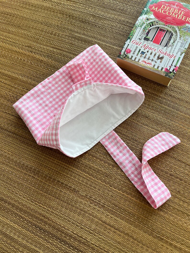 May include: A pink and white gingham bonnet with a white interior and long ties. The bonnet is next to a book with the title "G&uuml;l Ağacı Sokağı" by Debbie Macomber.