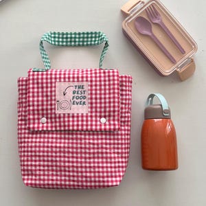 May include: A red and white gingham lunch bag with green gingham handles, a matching lunchbox with a fork and spoon, and an orange insulated water bottle. The lunch bag has a label that reads "THE BEST FOOD EVER."