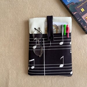 May include: A black fabric pouch featuring a white musical staff and note pattern with a cream-colored interior lining. The pouch has a central snap-button strap and contains a pair of round wire-rimmed glasses, three pens, and a small card.