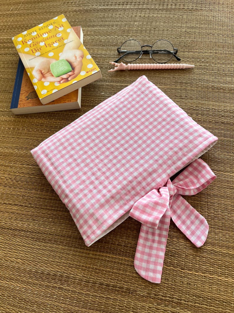 May include: A pink and white gingham pattern book sleeve with a matching bow. The image also shows a pair of round glasses, a pink stylus, and two books, one with the title "Kendime Ait G&ouml;r&uuml;nme Bir Işaret".